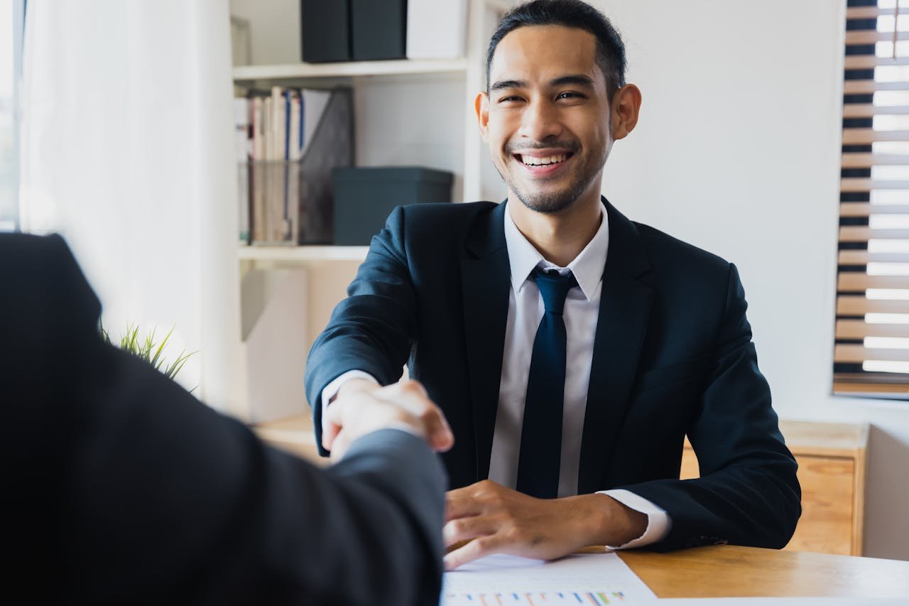 products-hero-img Confident businessman in suit shaking hands at office desk, symbolizing successful partnership.